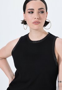 Czarny top na ramiączkach o ryflowanej strukturze, bez rękawów, z błyszczącego czarnego wykończenia wokół dekoltu i armhole. Złote kolczyki w kształcie kół podkreślają wygląd.