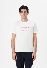 Weißes Baumwoll-T-Shirt mit kurzen Ärmeln, das auf der Vorderseite einen roten Grafikdruck mit "TOMMY HILFIGER NEW YORK CITY" und "Est. 1985" zeigt.