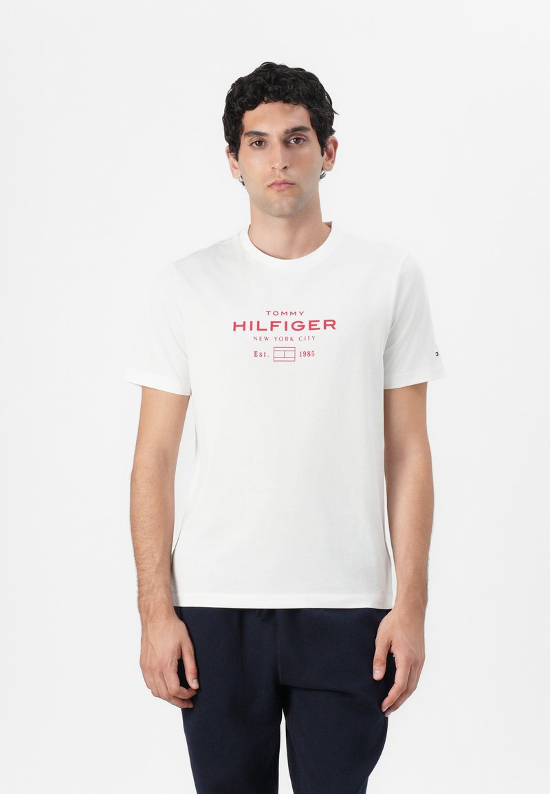 Fehér, rövid ujjú pamut póló, amelynek elején piros grafikával van díszítve: "TOMMY HILFIGER NEW YORK CITY" és "Est. 1985" felirattal.