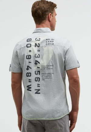Man wearing a light gray short-sleeve shirt with large black text and coordinates printed on the back, standing facing away.