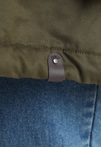 Veste vert olive à texture lisse, avec une lanière en cuir marron et un détail de bouton-pression, portée au-dessus d'un jean en denim bleu.