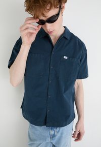 Dark navy short-sleeve shirt with two front pockets, button-down collar, and a small brand tag. Worn with light blue denim jeans.