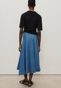 T-shirt noir à manches courtes associé à une longue jupe en denim bleue, dotée d'un design évasé et de coutures accentuées, portée avec des sandales noires.