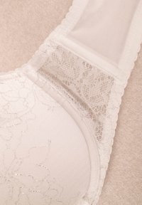 Sutien din dantelă albă cu modele florale, material neted pe cupe și un panou din plasă pe bretele. Marginile sunt crenelate pentru detaliu.