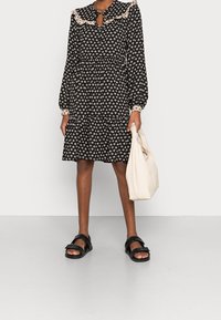 Robe noire avec motif floral crème, manches longues bouffantes, col orné de dentelle, décolleté en trou de serrure, ourlet à volants, associée à des sandales noires et un sac crème.