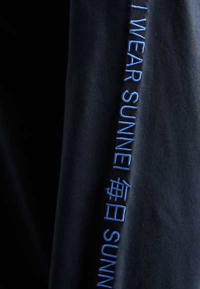 Must mustelari must tegija sinise tikandiga teksti "I WEAR SUNNEI" varrukal, lisaks hiina keeles märke.