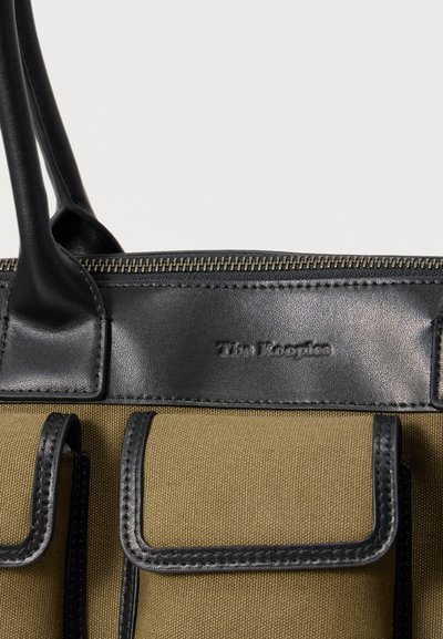 Κοντινή λήψη μιας καφέ υφασμάτινης τσάντας χειρός με μαύρες δερμάτινες λαβές, επένδυση και δύο μπροστινές τσέπες, με φερμουάρ κλεισίματος.