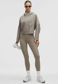 Kapat, grå vattentät jacka med huva och framfickor, tillsammans med beige leggings. Vita träningsskor och en vit vattenflaska.