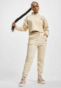 Beige kort sweatshirt med høy krage og glidelås, kombinert med matchende joggebukser med elastisk midje og sidelommer.