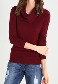 Maglione color bordeaux con collo a mantella in morbido tessuto lavorato a maglia, caratterizzato da maniche lunghe e polsini a coste. Abbinato a jeans di denim effetto usurato.