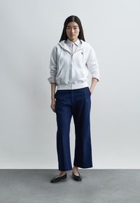 Νέα γυναίκα που στέκεται με το ένα χέρι στην τσέπη, φορά λευκή ζακέτα φούτερ με φερμουάρ, ριγέ πουκάμισο, ναυτικά παντελόνια με φαρδύ πόδι και μαύρα μοκασίνια, μπροστά σε απλό φόντο.