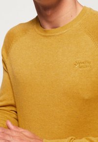 Senfgelbes Crewneck-Pullover aus weichem Stoff, mit einem dezenten Logo auf der Brust und Raglanärmeln.