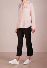 Blouse longue rose clair à manches longues avec un col en V, associée à un pantalon noir cropped et des baskets blanches. Tissu doux, coupe décontractée et fentes sur les côtés.