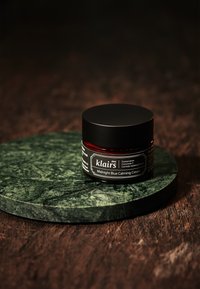 Svart sylinderformet krukke med skrulokk på en grønn marmoroverflate. Etiketten leser "klairs Midnight Blue Calming Cream." Kremen inni er synlig.