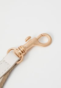 Chiusura in metallo dorato attaccata a una tracolla beige con cuciture visibili, caratterizzata da una texture liscia e bordi arrotondati.