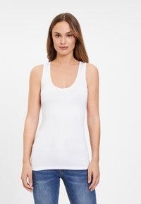 White tank top with a scoop neckline and wide straps, made of smooth, stretchy fabric. Paired with blue jeans.