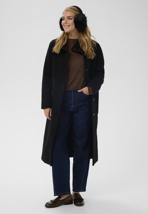 Abrigo largo negro con tela texturizada, suéter marrón debajo, jeans azules y mocasines marrones. Lleva orejeras de piel negra.