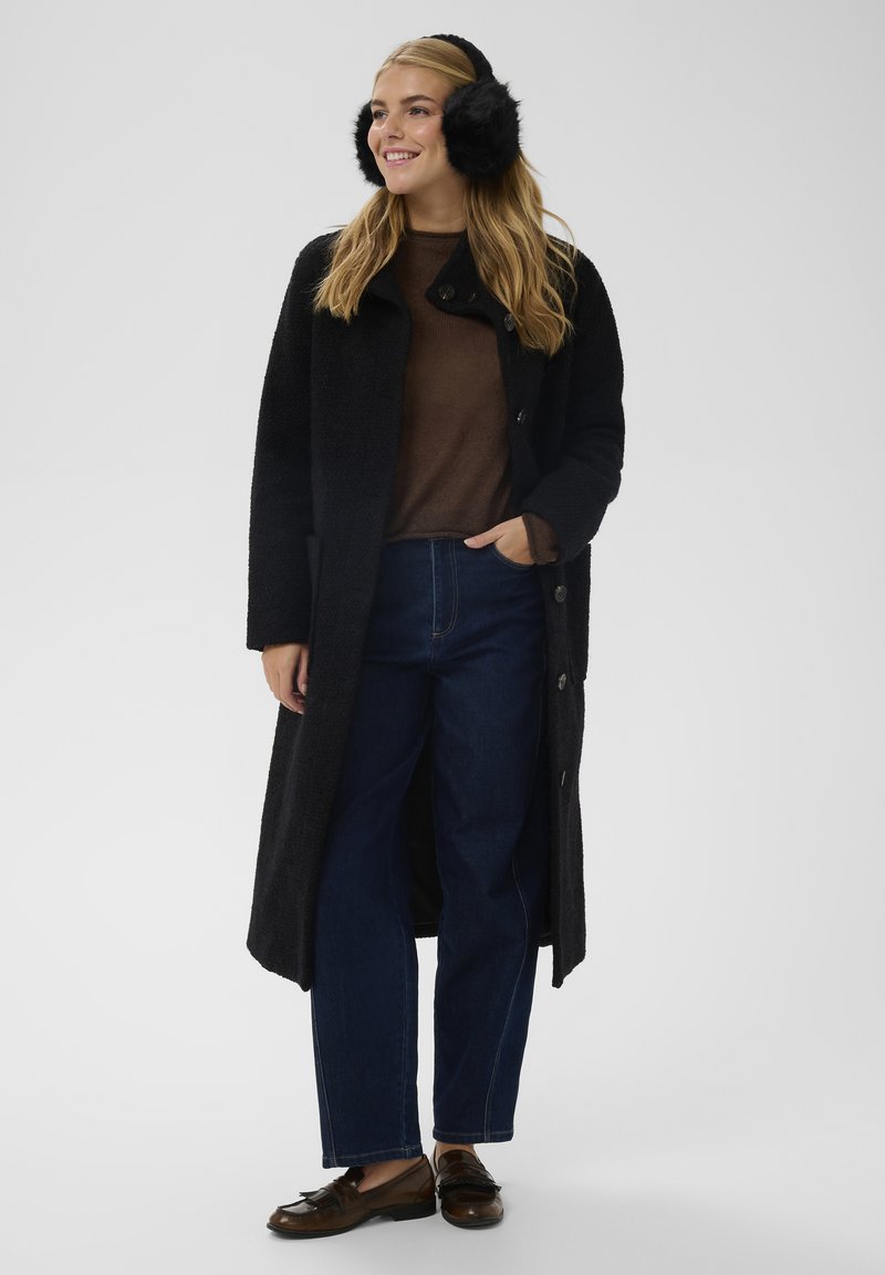 Dug kaput crne boje s teksturiranom tkaninom, ispod smeđi džemper, plave traperice i smeđe mokasinke. Ima crne krznene štitnike za uši.