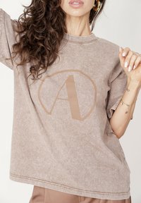 Brązowy, oversize'owy T-shirt wykonany z miękkiej tkaniny, z okrągłym logo przedstawiającym literę "A" w jaśniejszym odcieniu. Krótkie rękawy i luźny krój.