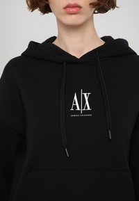 Personne portant un sweat à capuche noir avec le logo blanc « A|X Armani Exchange » centré sur la poitrine, la capuche reposant sur les épaules et les cordons pendants.