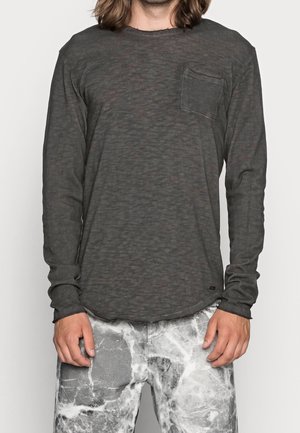 Hombre con camisa de manga larga gris oscuro con bolsillo en el pecho y pantalones con estampado de mármol blanco y gris, de pie con las manos relajadas a los lados.