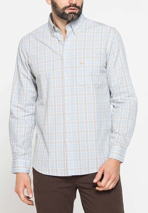 Uomo che indossa una camicia a quadri azzurra chiara e beige con bottoni e maniche lunghe e pantaloni marroni su sfondo bianco semplice.