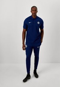 Mørkeblå sports-polo og bukser, lavet af glat stof. Indeholder Nike-logo og holdemblem på trøjen. Tæt pasform og tilspidsede bukser.