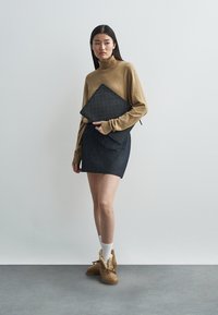 Custodia per laptop a quadri neri tenuta contro un maglione a collo alto beige, abbinata a una gonna di denim scuro e scarpe slip-on marroni con dettagli in pelliccia.