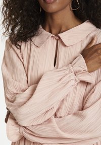 Blusa rosa pallido in tessuto strutturato e arricciato; presenta un colletto rotondo, chiusura con bottoni e polsini arricciati su maniche lunghe.