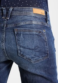 Vaqueros de mezclilla en azul oscuro con un bolsillo trasero que incluye una etiqueta de marca de cuero. La tela presenta un desvanecimiento sutil y una textura suave.