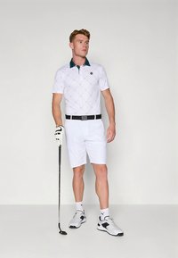 Vit golfpolo med grön krage och diamantmönster, kombinerad med vita shorts. Utrustad med svart bälte och golfhandske, håller i en klubba.