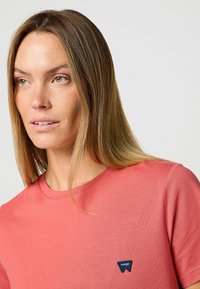Korallefarbiges T-Shirt aus weicher Baumwolle, mit rundem Halsausschnitt und kurzen Ärmeln, versehen mit einem blauen dreieckigen Logo auf der linken Brust.