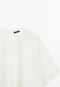 Chemise blanche à manches courtes avec un col rond et un petit détail de bouton à l'arrière, dotée d'une poche et d'un design minimaliste.