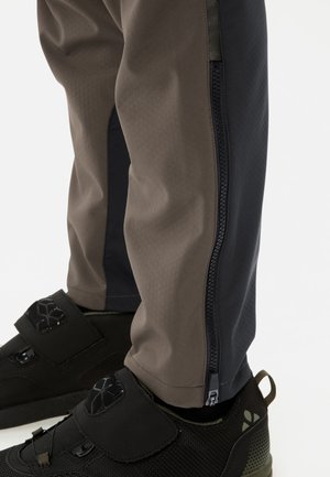 Untere Beine in braunen und schwarzen Hosen mit seitlichen Reißverschlüssen, kombiniert mit schwarzen Sportschuhen mit Klettverschlüssen.