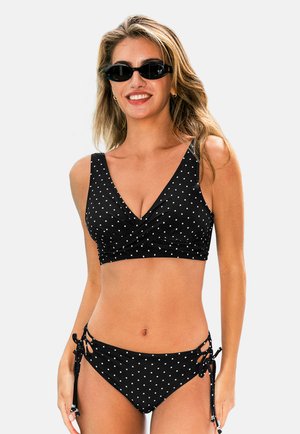 Uśmiechnięta kobieta w czarno-białym bikini w groszki z bocznymi wiązaniami i czarnymi okularami przeciwsłonecznymi, stojąca na jednolitym tle.