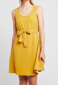 Een mouwloos A-lijn jurkje van mosterdgele stof, met een knoopriem in de taille en een uitlopende zoom met een asymmetrische snit.