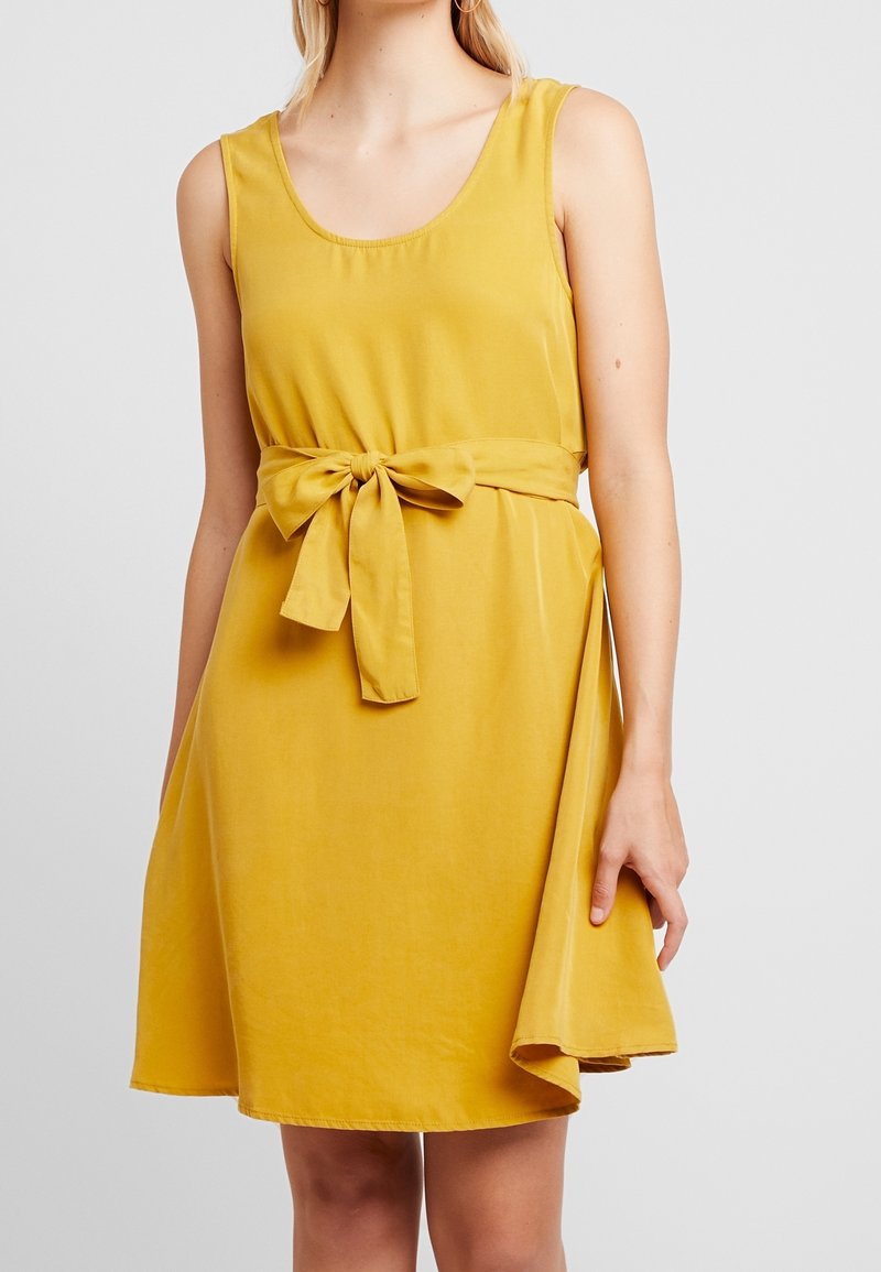 Een mouwloos A-lijn jurkje van mosterdgele stof, met een knoopriem in de taille en een uitlopende zoom met een asymmetrische snit.