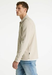 Chaqueta beige con textura, diseño con cuello, cierres de botones y dobladillo ligeramente curvado. Combinada con vaqueros de mezclilla azules.