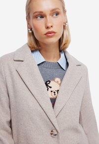 Beige wollen blendjas met geknipt revers, enkele knoopsluiting, zachte textuur, gedragen over een grijze trui met een teddybeer ontwerp en een blauw gestreepte shirt.