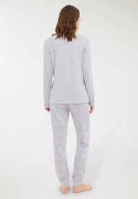 Donna in piedi a piedi nudi, girata di spalle, indossa una maglietta a maniche lunghe grigia chiara e pantaloni da pigiama abbinati con motivi di fiocchi rosa.