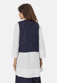 Jeansweste mit einer dunkelblauen Waschung, ärmellosem Design und einfarbiger Optik. Getragen über einem weißen, langärmligen Hemd mit detaillierten Manschetten.