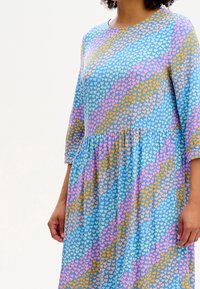 Žena v modrých šatech se nařaseným pasem a mnohobarevným srdíčkovým vzorem v růžové, žluté a světle modré.