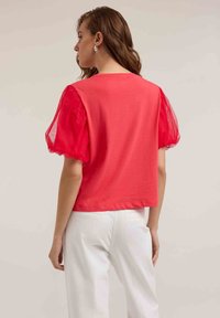 Top rojo de manga corta con mangas abullonadas transparentes; presenta una textura suave y un corte relajado, combinado con pantalones blancos de cintura alta.