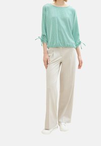 Žena oblečená v mátově zelené blůze s uvázanými 3/4 rukávy, béžových širokých kalhotách a bílých teniskách, stojící na jednobarevném pozadí.