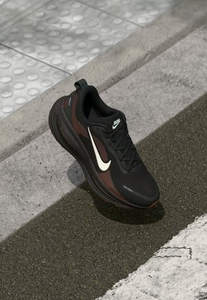 Chaussure de course Nike noire et marron avec un swoosh blanc sur un revêtement texturé près d’un pavage tactile et d’un trottoir.