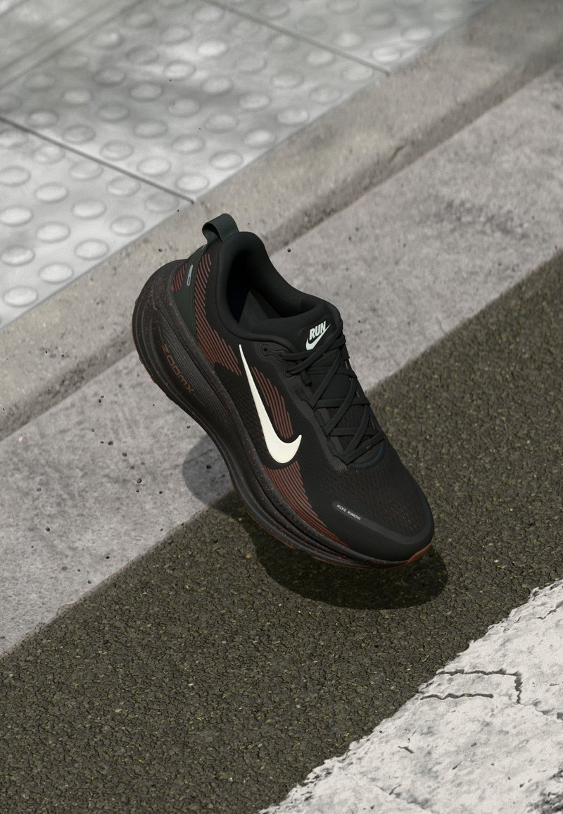 Chaussure de course Nike noire et marron avec un swoosh blanc sur un revêtement texturé près d’un pavage tactile et d’un trottoir.