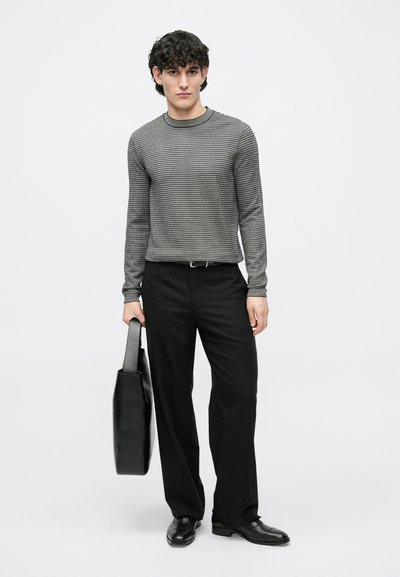 Szürke, texturált kötött pulóver hosszú ujjakkal, fekete nadrággal és fekete cipővel párosítva. Egy fekete táskát tart a kezében.