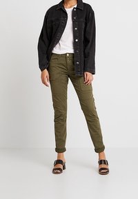 Svart denimjacka över en vit t-shirt, tillsammans med olivgröna cargobyxor med fickor. Svarta sandaler med remmar fulländar looken.