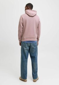 Helle rosa Kapuzenjacke mit Kordelzug, kombiniert mit lockeren blauen Jeans und beigen Schuhen. Die Textur wirkt weich und der Schnitt ist entspannt.