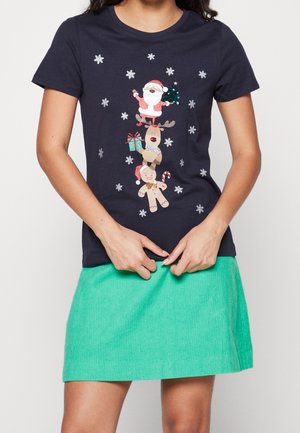 Kobieta w granatowym t-shircie z układem Świętego Mikołaja, renifera i piernikowego ludzika otoczonych płatkami śniegu oraz zieloną spódnicą.